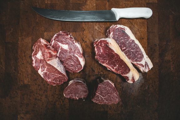 Viande crue de différentes coupes avec un grand couteau sur une planche en bois.